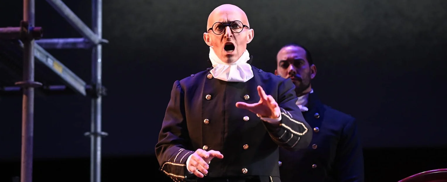 Corey Trahan, dressed in a period costume with glasses, signs on stage