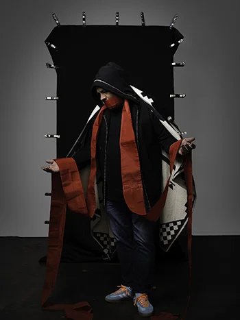 A figure dressed in a ceremonial cloak of mourning stands in front of a black drapery evocative of a doorway