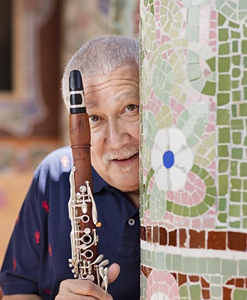 Paquito D'Rivera peeks from behind a tiled column, holding a rosewood clarinet