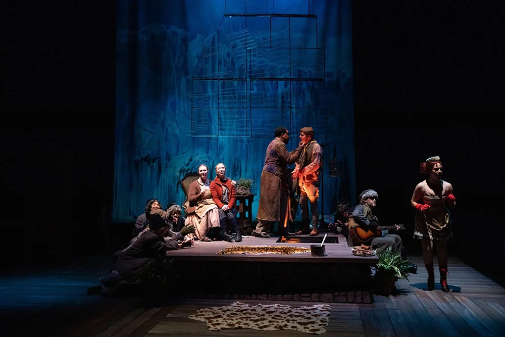 A small group of stage performers, illuminated by a strong column of light, are surrounded by darkness